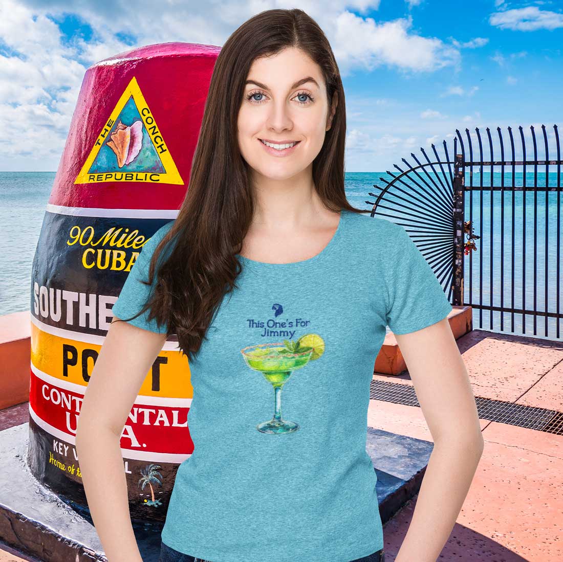 Woman wearing a blue t-shirt with a cocktail graphic, standing in front of a colorful bottle and ocean view.