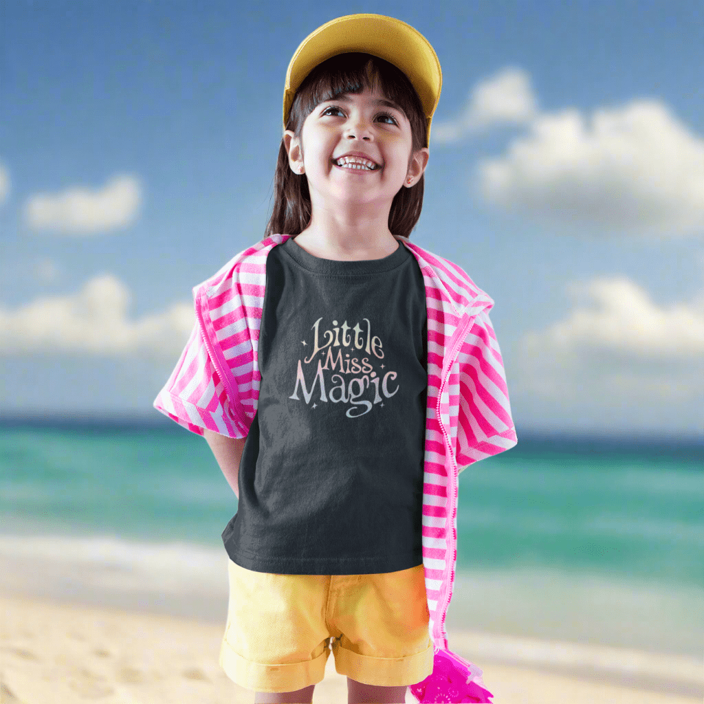 Child wearing a 'Little Miss Magic' shirt with a pink cape and yellow shorts on St. Pete Beach