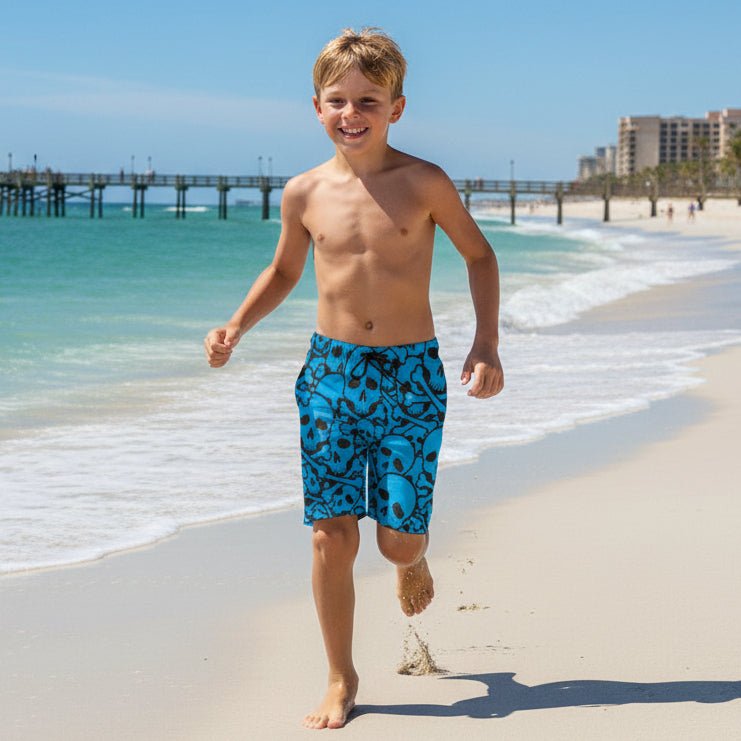 Young boy in blue pirate themed swim Skull & Bones Beach Shorts running on a sandy beach with ocean and pier in the background