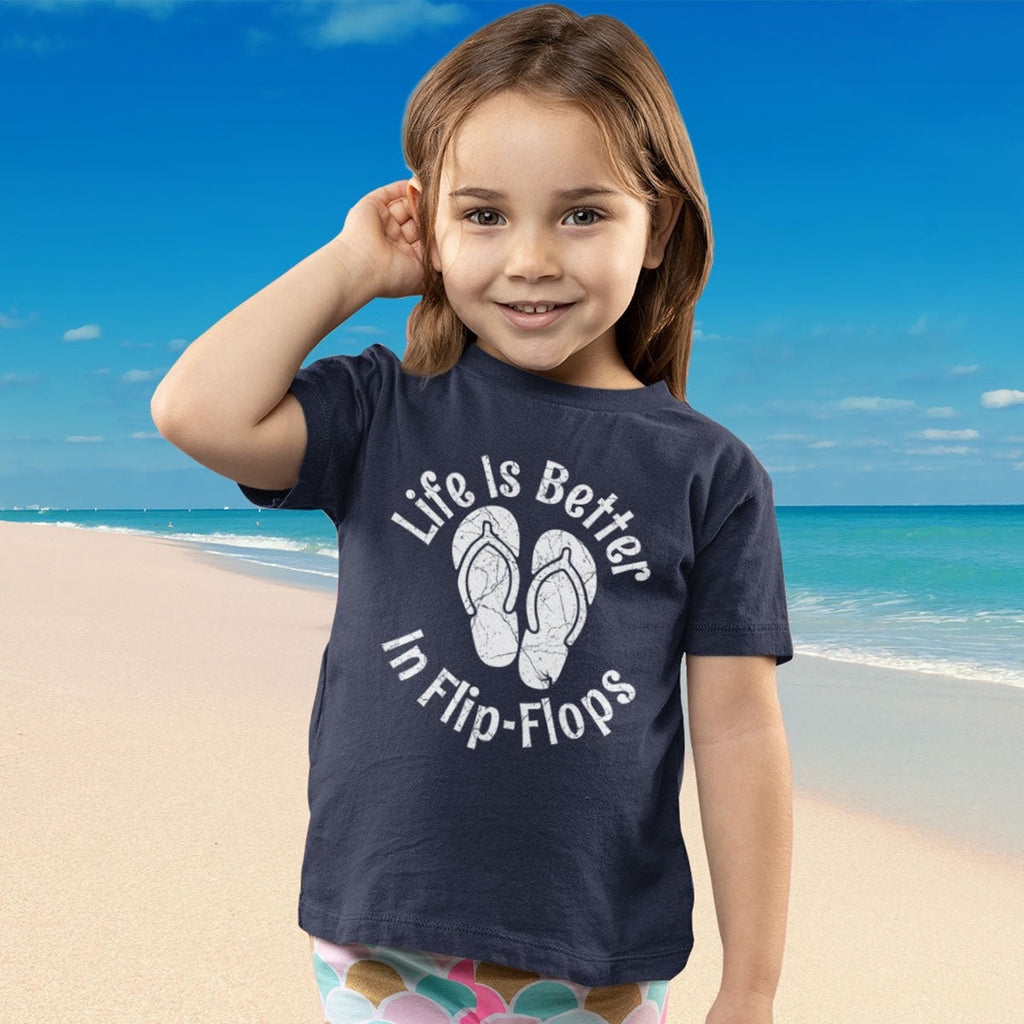 Child wearing a navy blue Flip-Flop Toddler Tee with 'Life is Better in Flip-Flops' text on a beach background