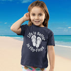 Child wearing a navy blue Flip-Flop Toddler Tee with 'Life is Better in Flip-Flops' text on a beach background