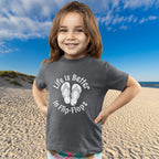 Child wearing a t-shirt with 'Life is Better in Flip-Flops' text on a beach
