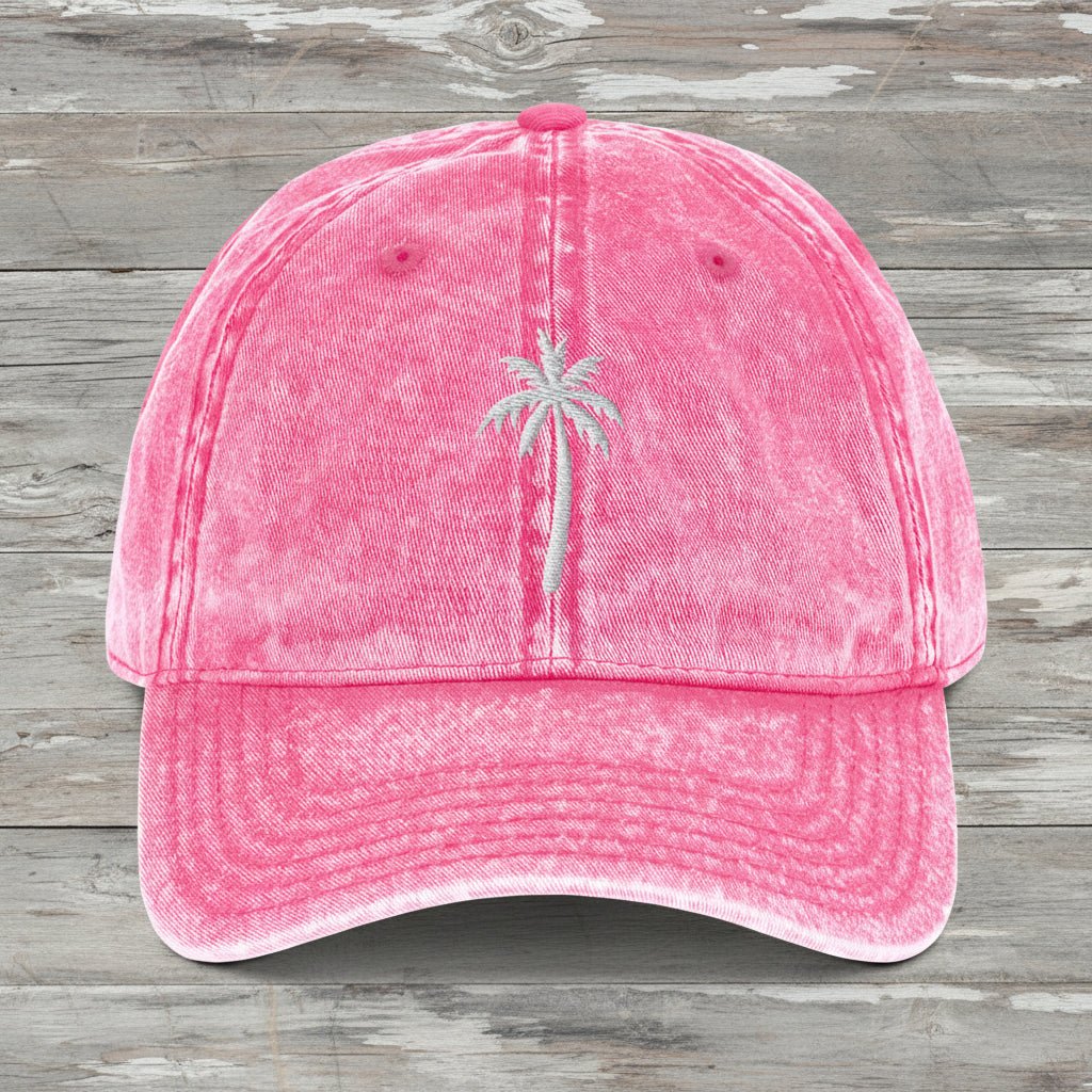 A pink acid-wash cotton baseball cap featuring a small embroidered palm tree on the front, set against a rustic wood background.