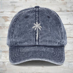 A washed blue denim baseball cap featuring a white embroidered palm tree on the front, set against a rustic wood background