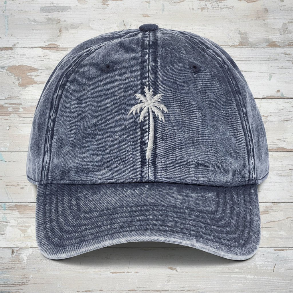 A washed blue denim baseball cap featuring a white embroidered palm tree on the front, set against a rustic wood background