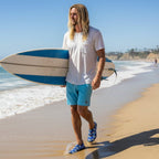 Surfer dude with long stringy blond hair wearing shaka slippers walking along the water on La Jolla Shores Beach with a surfboard