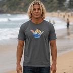A man with blonde hair stands on a sandy beach wearing a dark gray Shaka Wave T-Shirt with a colorful shaka hand graphic on the chest, paired with dark blue jeans.
