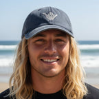A smiling man with long blonde hair wearing a dark blue baseball cap with a white hand gesture logo, photographed outdoors on a beach with the ocean in the background.