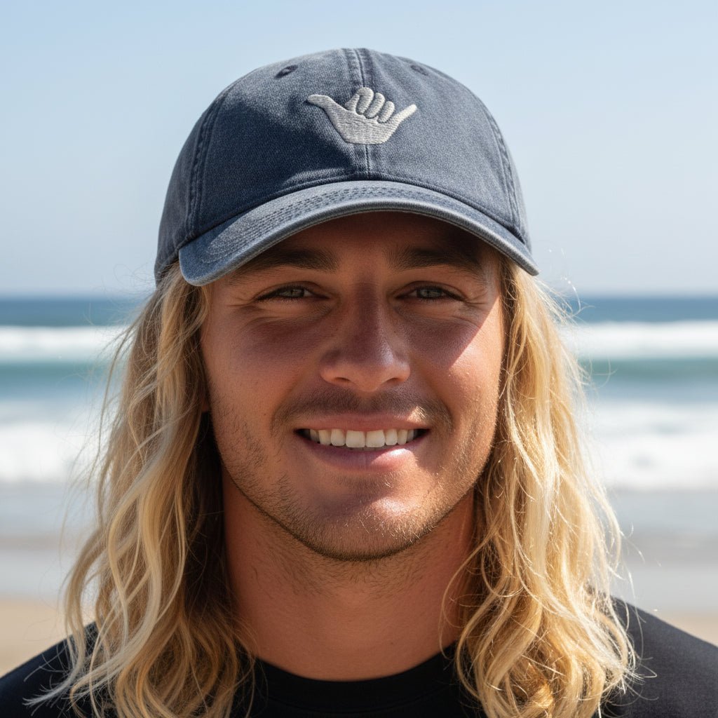 A smiling man with long blonde hair wearing a dark blue baseball cap with a white hand gesture logo, photographed outdoors on a beach with the ocean in the background.