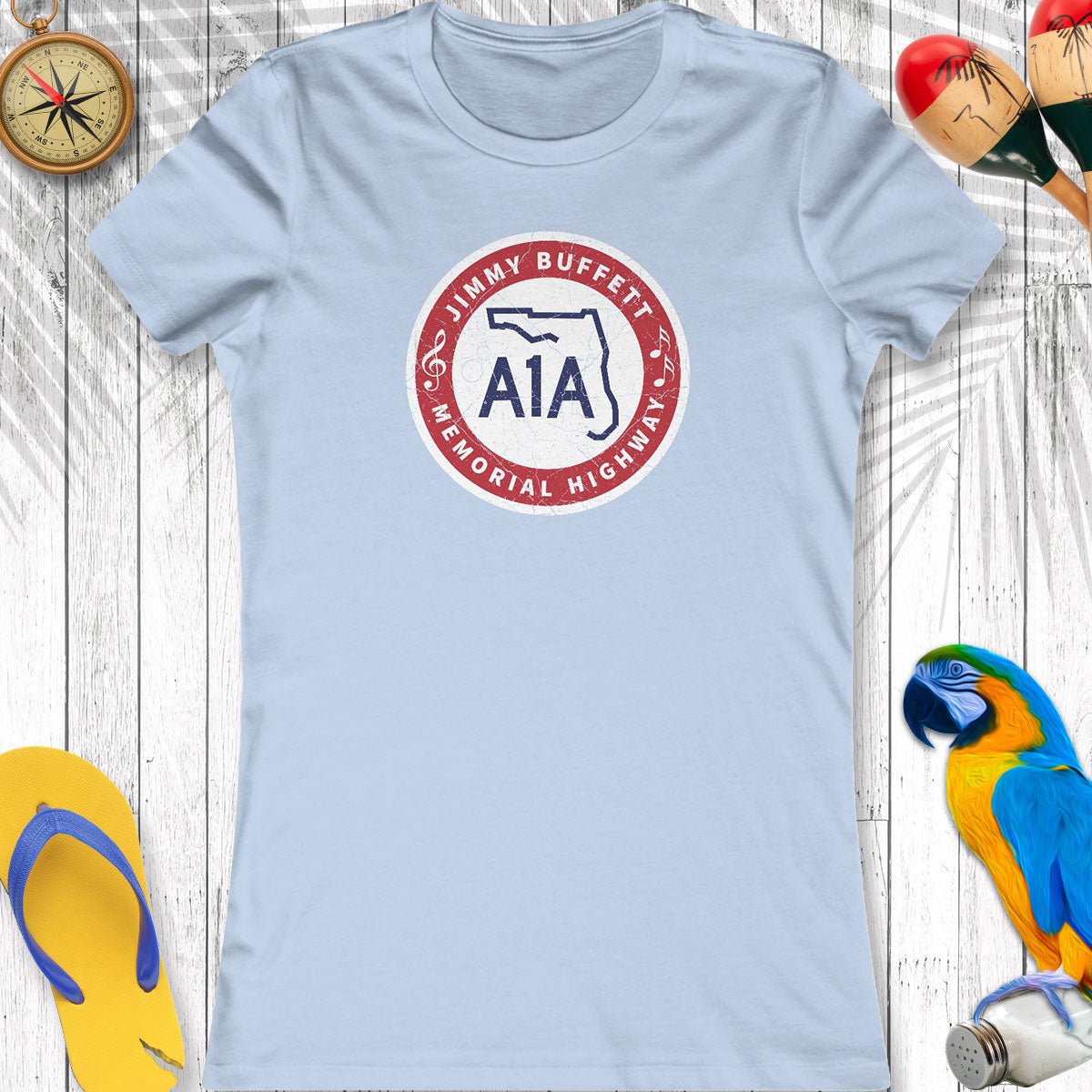 Light blue t-shirt with a circular logo featuring 'A1A' and 'Jimmy Buffett Memorial Highway' on a wooden surface.
