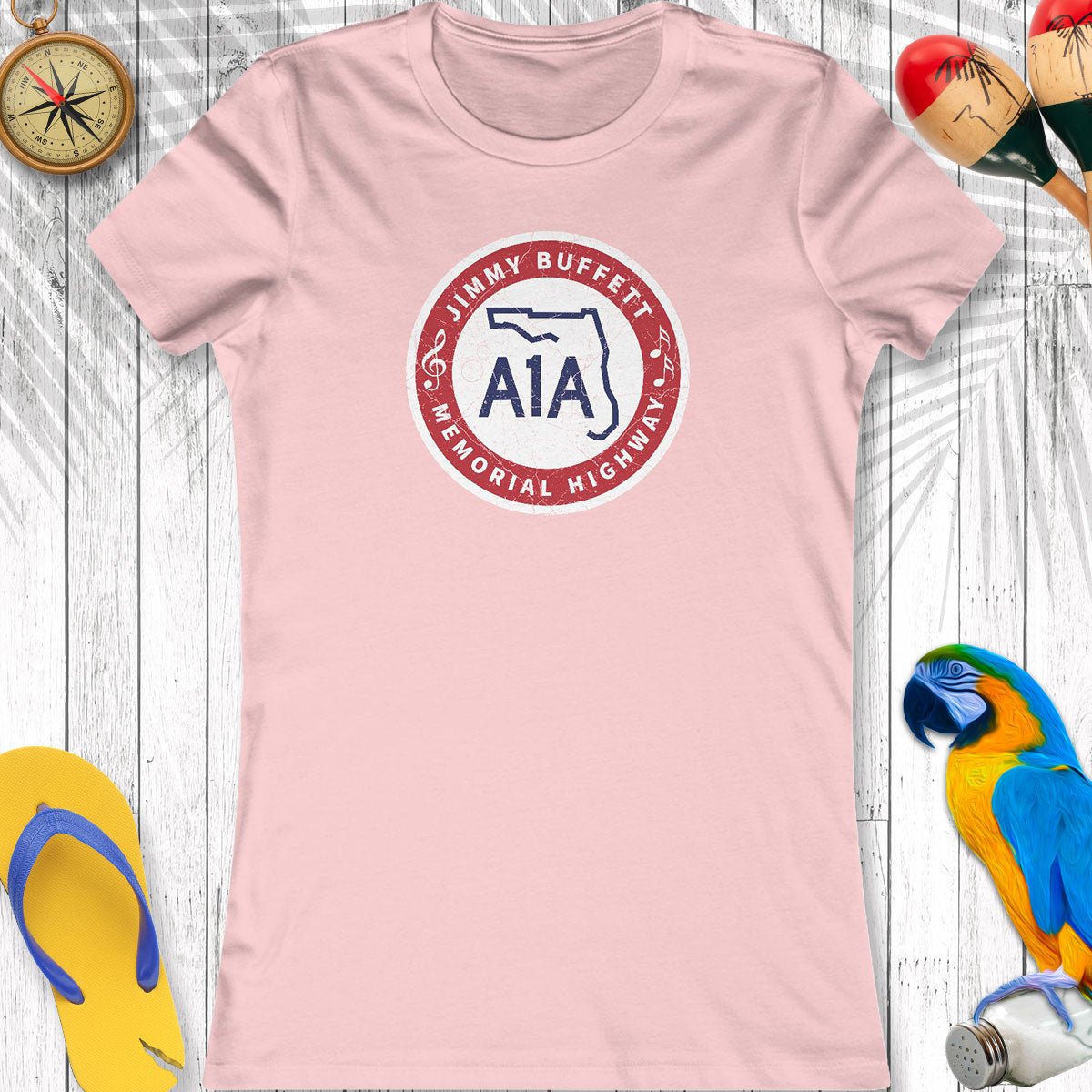 Pink t-shirt with a logo and text on a wooden surface with beach accessories.