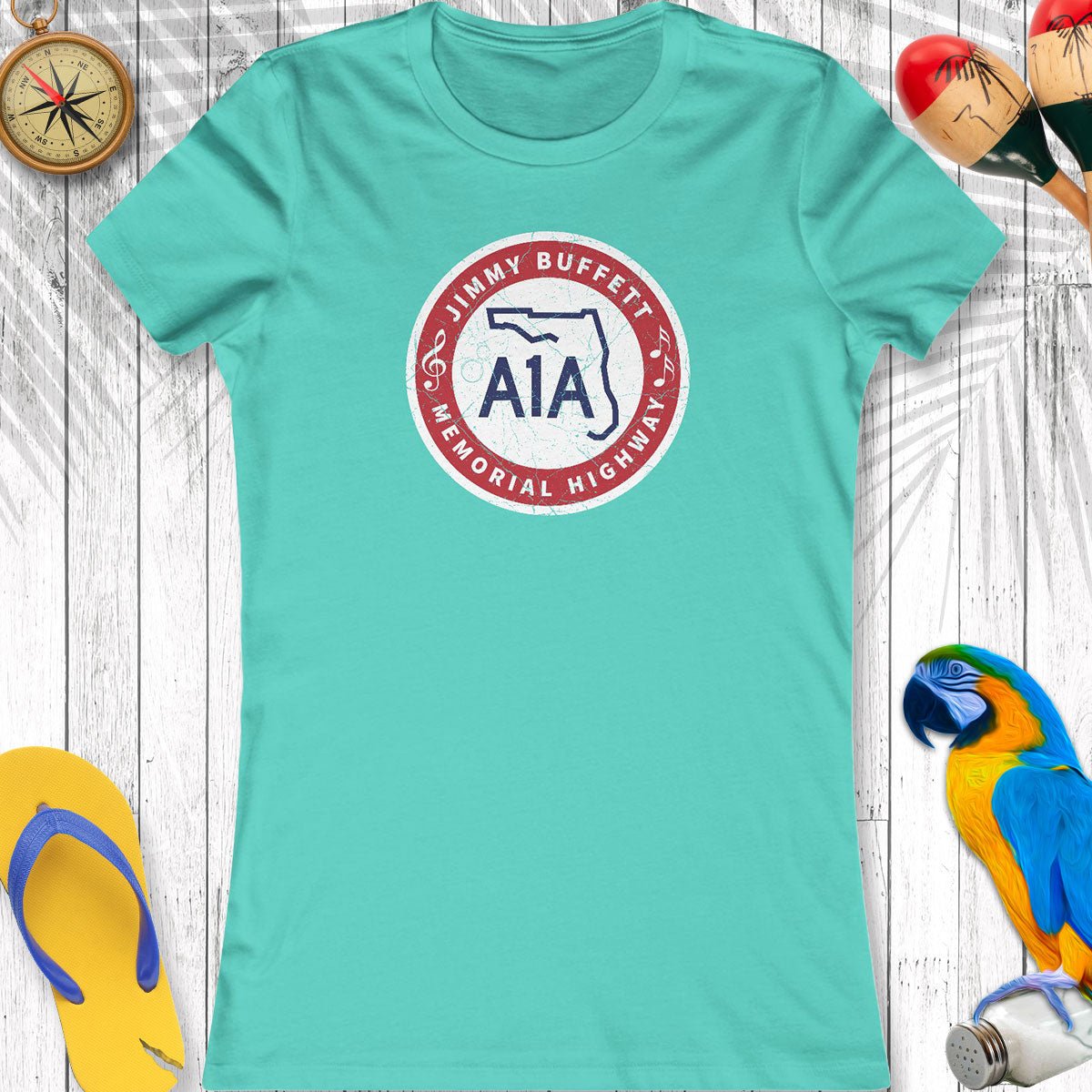 Teal t-shirt with a circular logo featuring 'Jimmy Buffett A1A Memorial Highway' on a wooden surface with beach accessories.