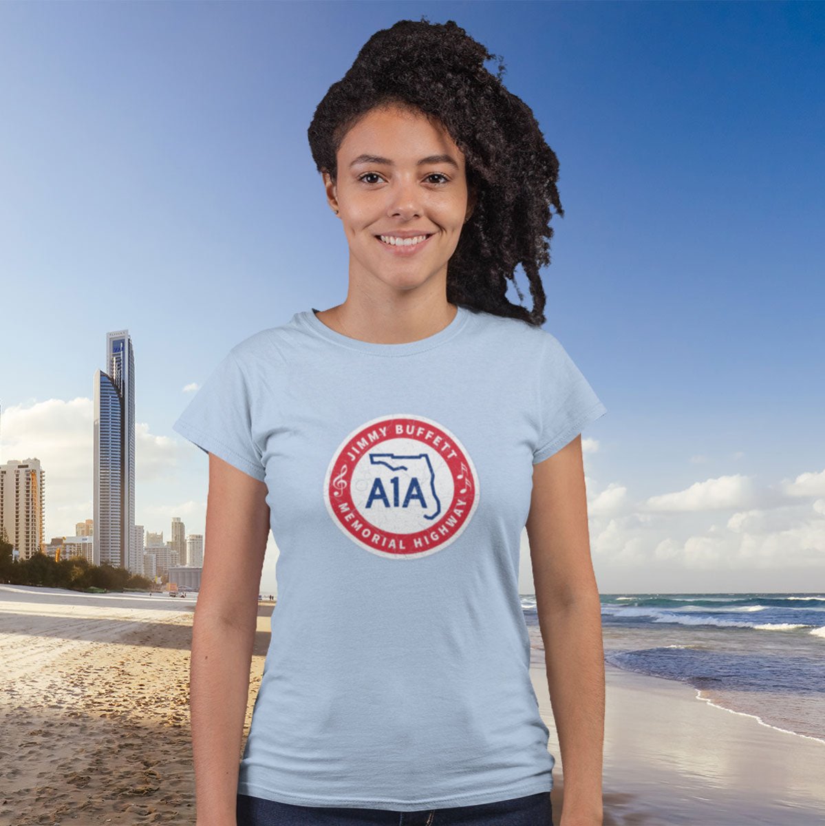 Person wearing a light blue t-shirt with a logo on a beach with city skyline in the background