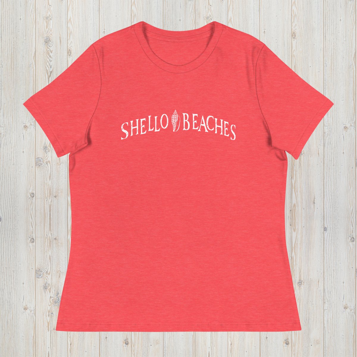 A heather coral red short-sleeve t-shirt with 'SHELLO BEACHES' printed in white across the chest, laid flat on a light wood plank background.