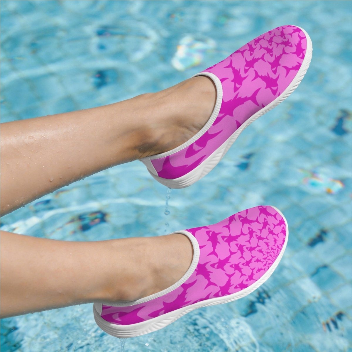A pair of pink slip-on water shoes with a darker pink shark pattern and white soles, shown being worn with a person's legs visible above a swimming pool.