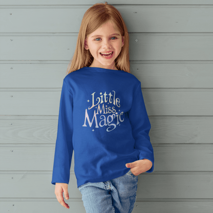 Child wearing a blue sweatshirt with 'Little Miss Magic' text against a gray wooden wall.