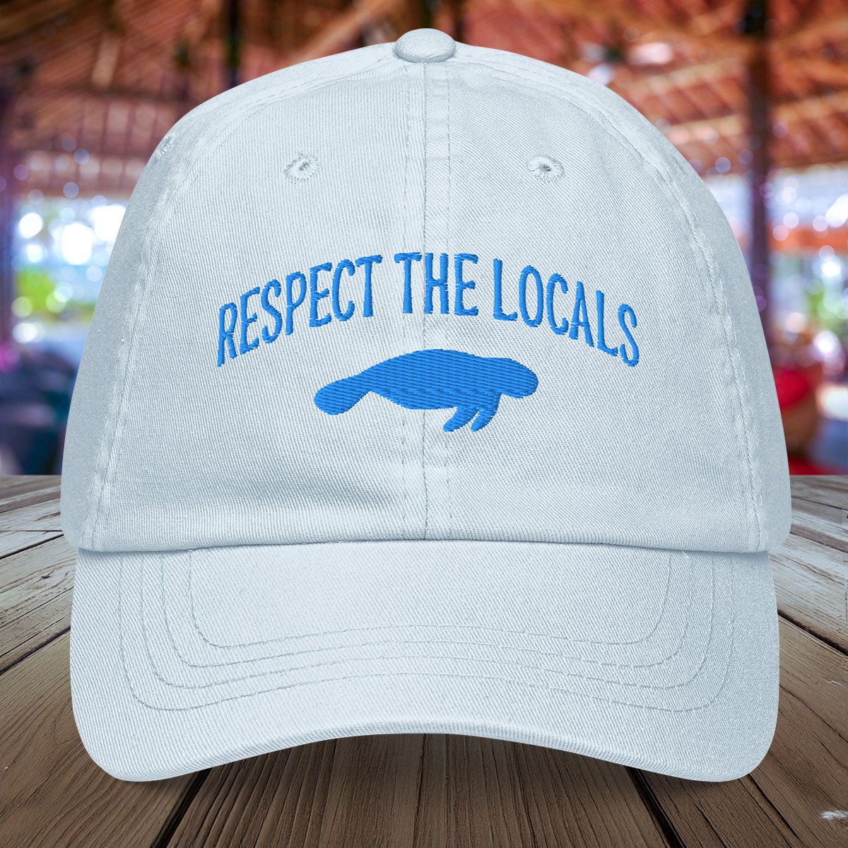 Pastel light blue Manatee cap with 'Respect the Locals' text and blue graphic on a wooden surface
