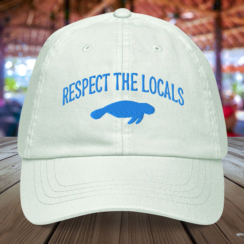 Pastel mint green Manatee cap with 'Respect the Locals' text and blue embroidered manatee on a wooden surface.