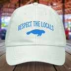 Pastel mint green Manatee cap with 'Respect the Locals' text and blue embroidered manatee on a wooden surface.