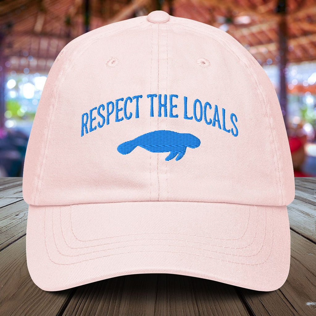 Pastel Pink Manatee cap with 'Respect the Locals' text and blue manatee silhouette on a wooden surface.