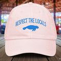 Pastel Pink Manatee cap with 'Respect the Locals' text and blue manatee silhouette on a wooden surface.