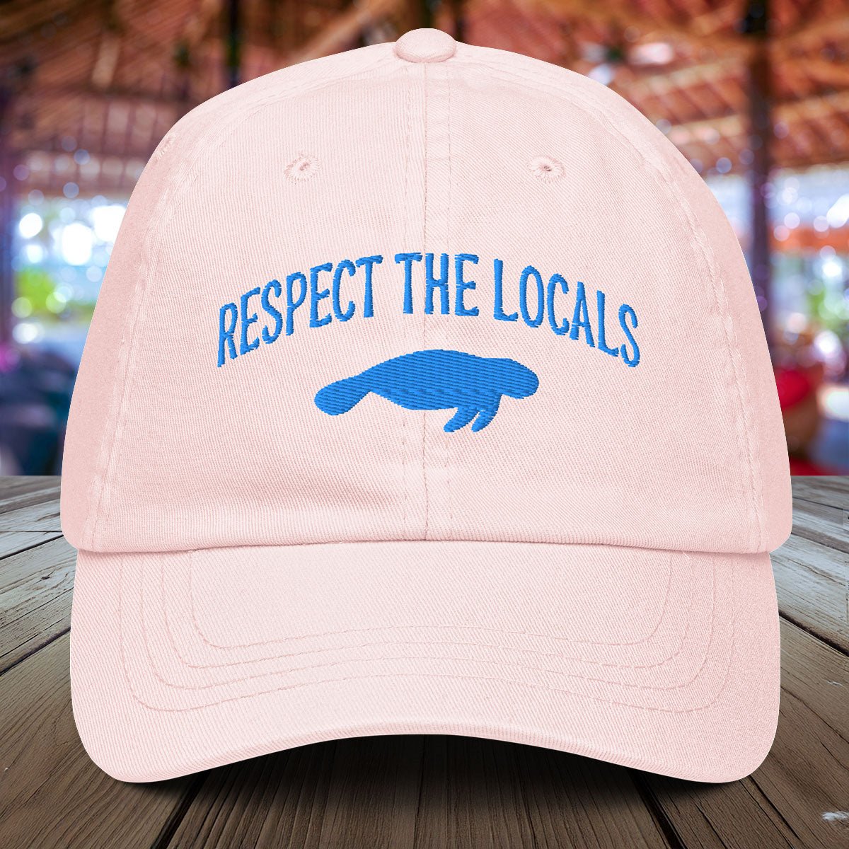 Pastel Pink Manatee cap with 'Respect the Locals' text and blue manatee silhouette on a wooden surface.