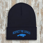 A navy blue ribbed knit beanie hat with a folded cuff, featuring embroidered blue text reading 'RESPECT THE LOCALS' above a blue manatee graphic, shown on a rustic wood background.