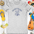 Gray t-shirt with a cocktail graphic and text, surrounded by flip-flops, a parrot, and a ukulele on a wooden background.