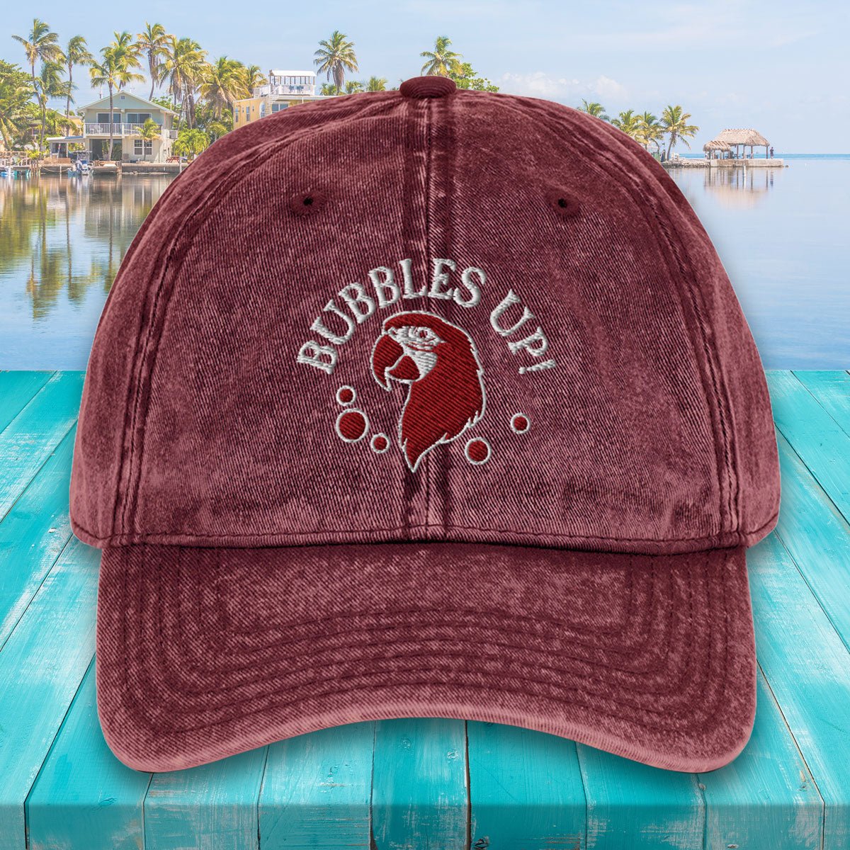 Maroon cap with 'Bubbles Up!' design on a turquoise surface with a pool and palm trees in the background