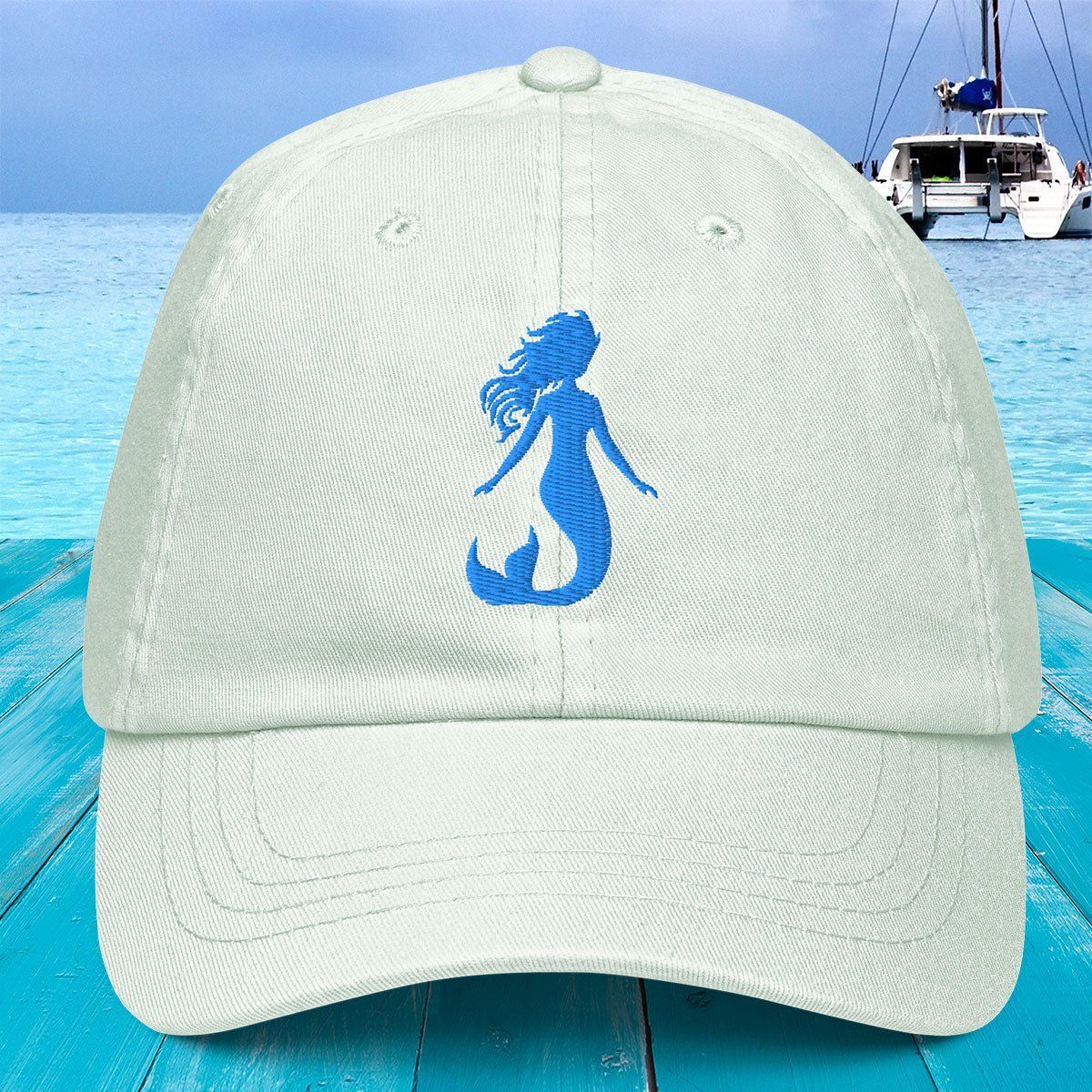 Beige cap with blue mermaid silhouette on a blue wooden surface with a boat in the background
