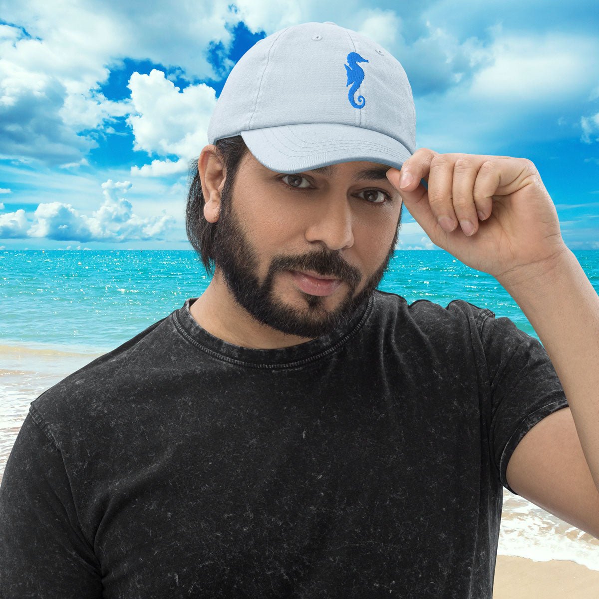 Man wearing a pastel light blue cap with a blue embroidered seahorse on a beach