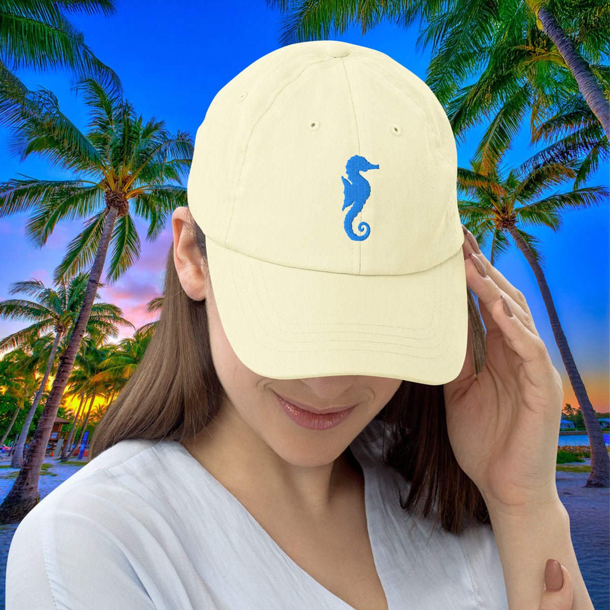 Person wearing a pastel yellow cap with a blue seahorse design against a tropical background with palm trees and sunset.