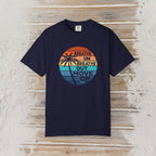 A navy blue short-sleeve t-shirt with a retro circular graphic on the chest, laid flat on a light-colored wooden plank background. The graphic features a sunset design with palm trees and the text 'BREATHE IN BREATHE OUT MOVE ON'.