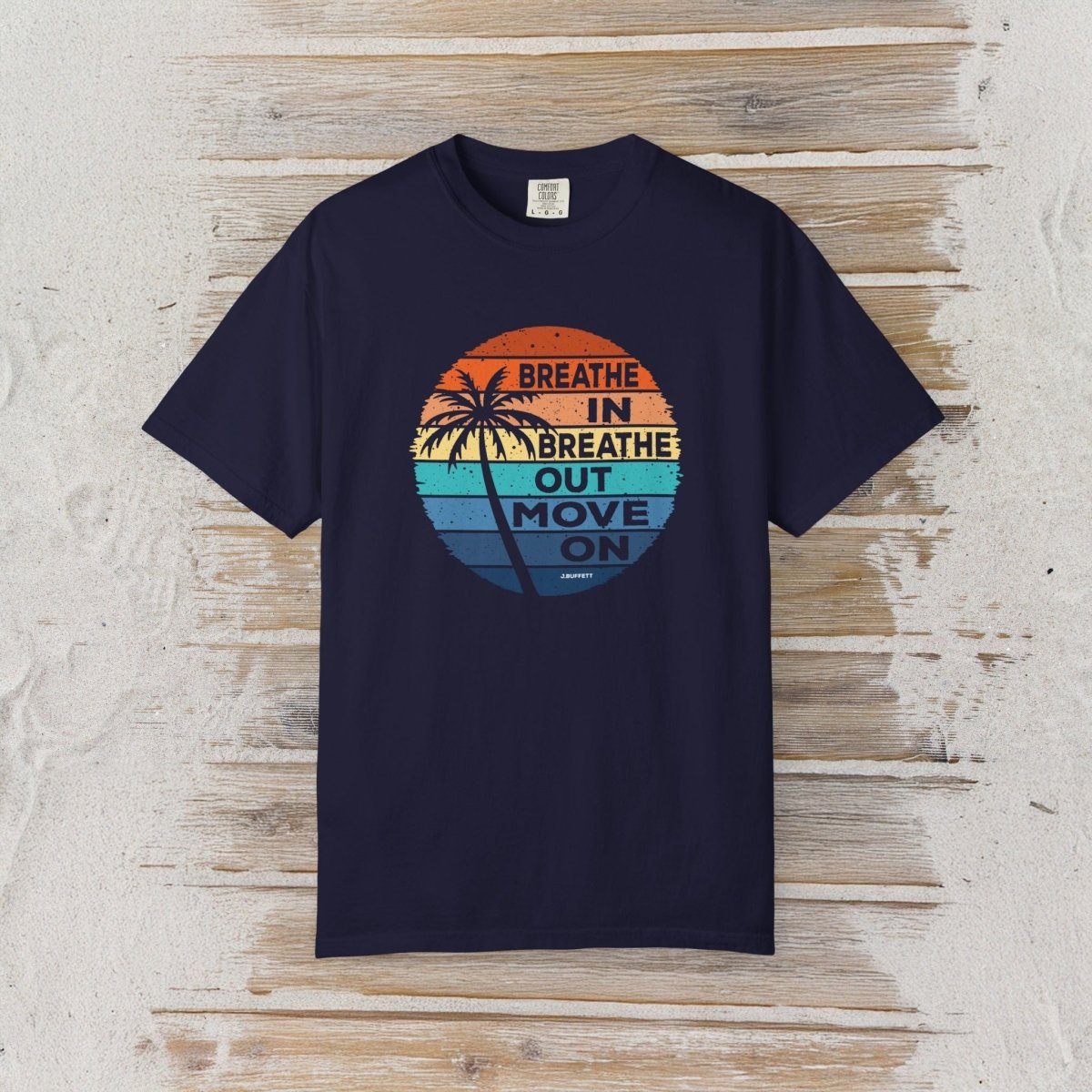 A navy blue short-sleeve t-shirt with a retro circular graphic on the chest, laid flat on a light-colored wooden plank background. The graphic features a sunset design with palm trees and the text 'BREATHE IN BREATHE OUT MOVE ON'.