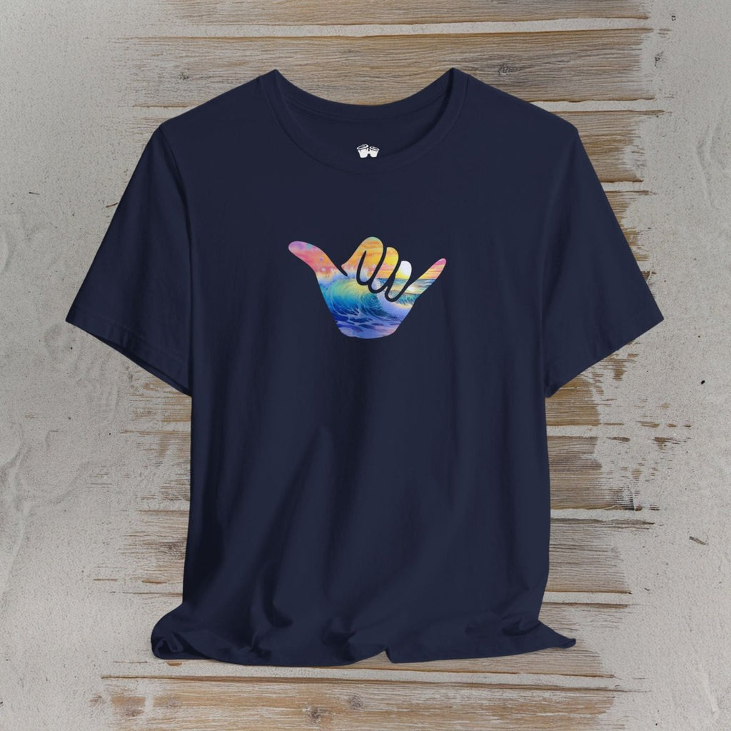 A navy blue short-sleeve t-shirt with a colorful Hawaiian 'shaka' hand graphic on the chest, displayed flat against a rustic light wood background.