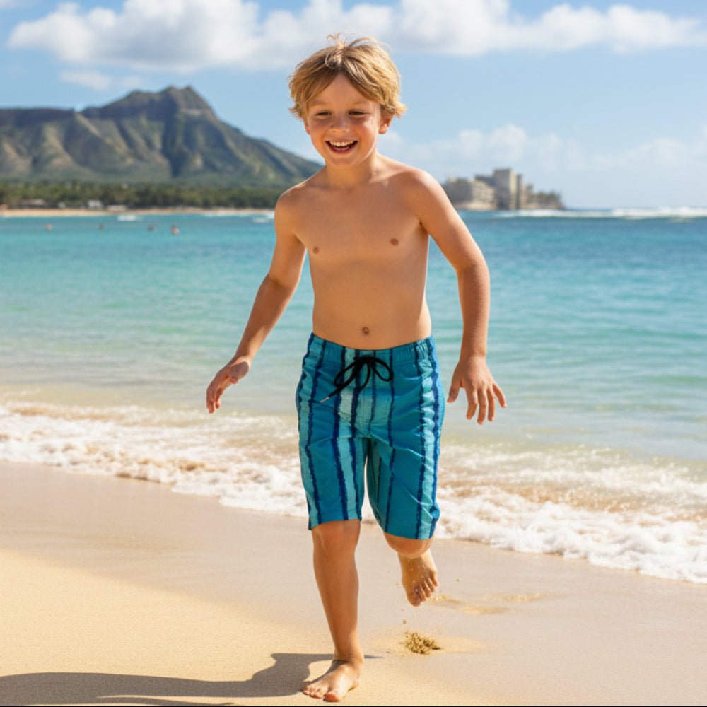 Child in aqua blue striped Board Shorts standing on Waikiki beach with surf and Diamond Head mountain in the background.