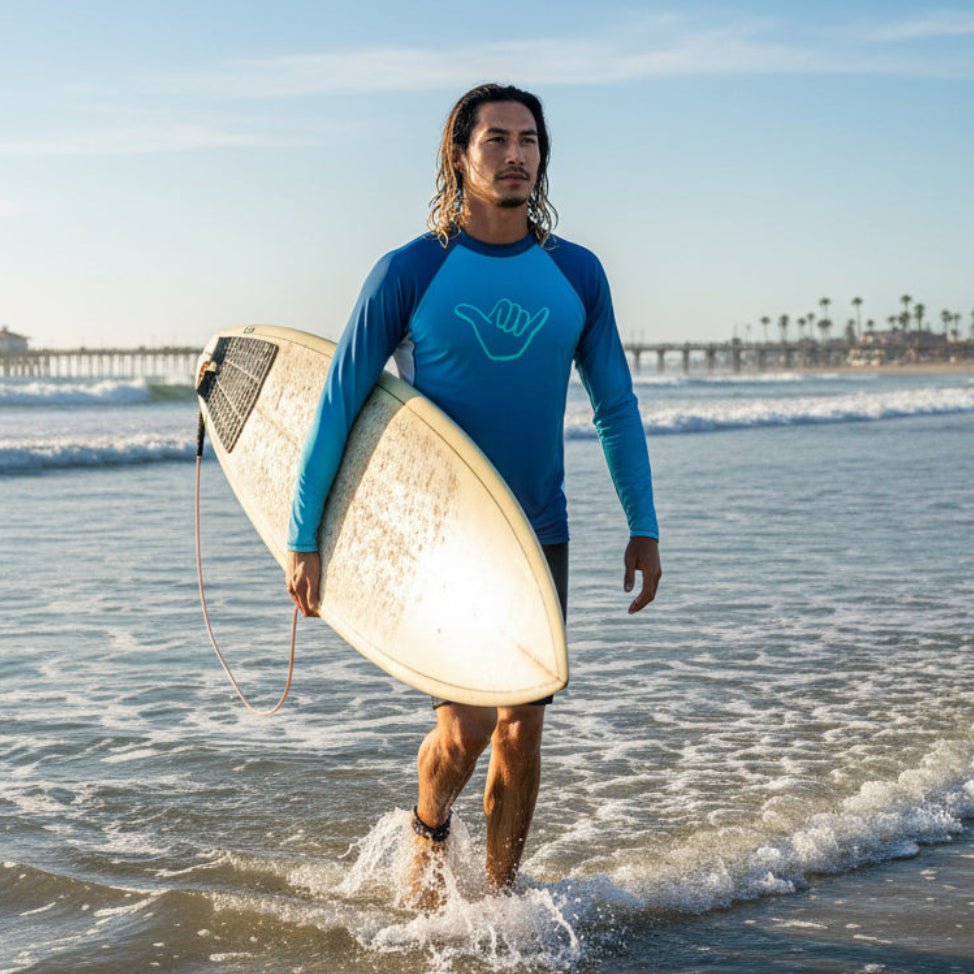 Surfer dude with long black hair walking through the waves at Oceanside Beach California, holding a surfboard and wearing Blue and navy long-sleeve rashie with a green shaka hand icon
