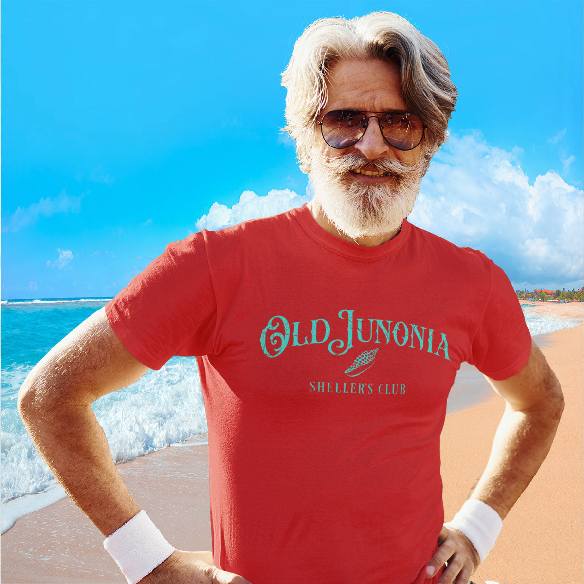 Man wearing a red 'Old Junonia' t-shirt on a beach with blue sky and ocean.