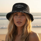 A black denim bucket hat with a distressed frayed edge and a white embroidered 'Old Sheller' over a junonia seashell on Anna Maria Island Beach