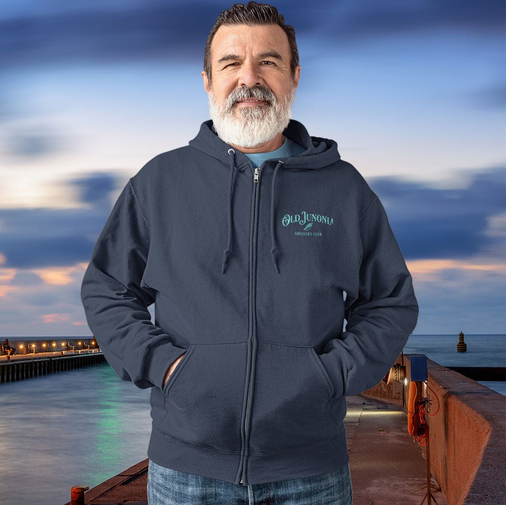 Man wearing a dark hoodie with a logo standing on a dock with a scenic background