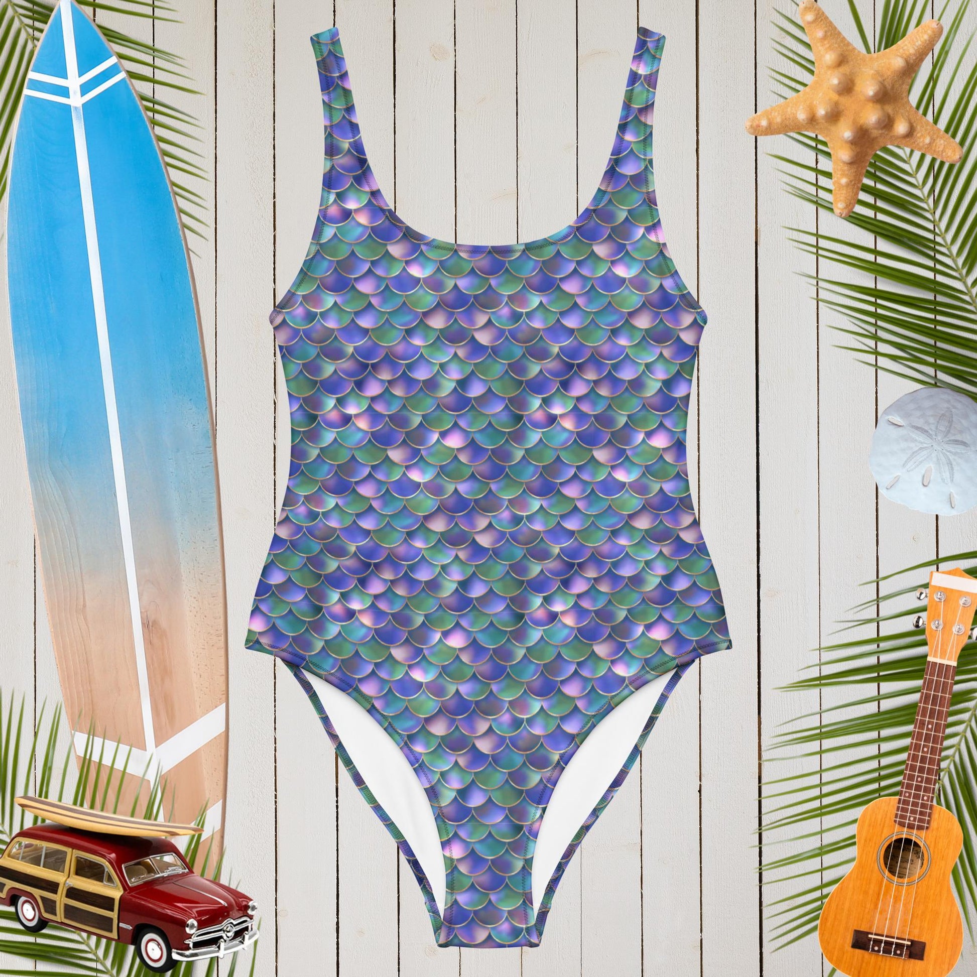 One Piece Mermaid Swimsuit on a rustic wood wall next to a surfboard and a Ukulele. 