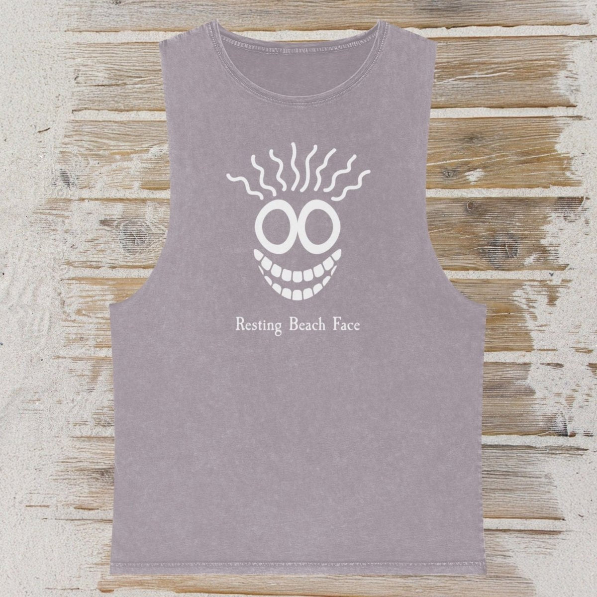 A sleeveless heather gray tank top featuring a white graphic of a skull with wavy hair and the text 'Resting Beach Face' on the front, displayed flat on a rustic wooden plank background.