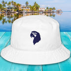 White bucket hat with a blue logo on a turquoise wooden surface with a scenic background of houses and palm trees.