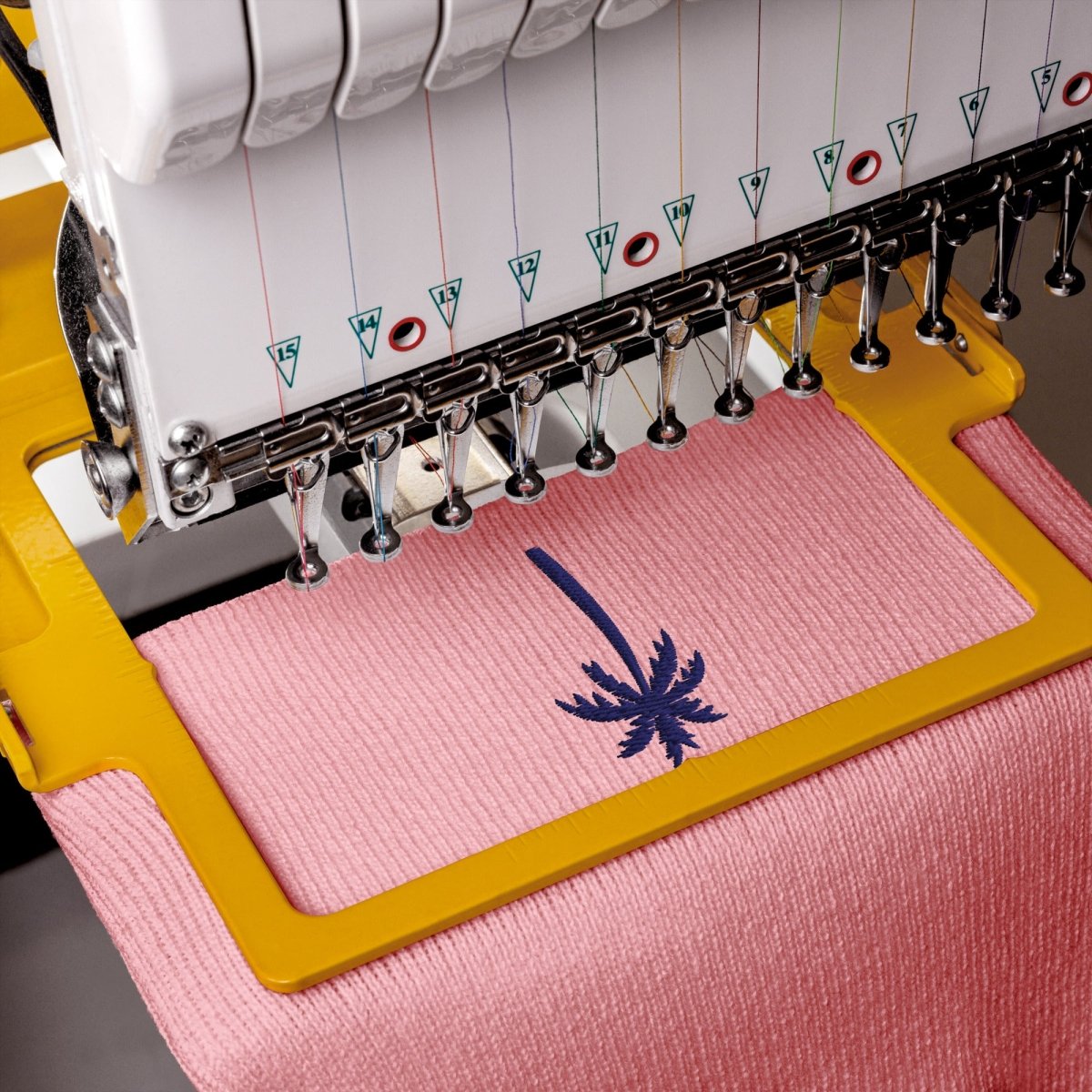 Close-up of an embroidery machine stitching a pink fabric piece with a dark blue palm tree design. The machine has a white body with multiple embroidery threads and a yellow frame holding the fabric.