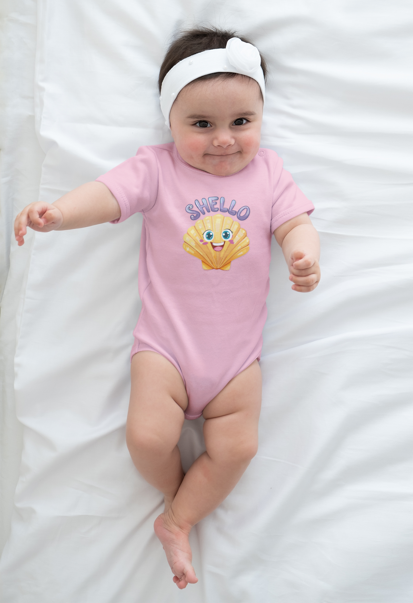 Baby wearing a pink onesie with a shell design and 'SHello' text on a white blanket