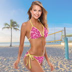 Woman in a pink and yellow bikini on a beach with palm trees and a volleyball net in the background.