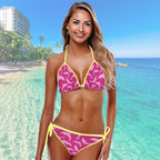 Bikini model in a pink and yellow bathin suit standing on a beach with clear blue water and palm trees in the background.