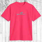 A bright pink short-sleeve t-shirt with a small gray silhouette of a cat on a surfboard across the chest, displayed flat against a white wood plank background.