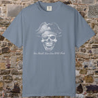 A slate blue crewneck t-shirt featuring a white graphic of a skull wearing a pirate hat with a skull-and-crossbones symbol, and the text 'You Should Never Wish Trust' printed below. The t-shirt is displayed flat against a rustic, textured stone brick wall background.