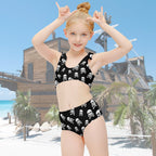 Child wearing a black and white skull patterned two-piece swimsuit on beach near a ship shaped building